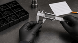 Gloved hands measuring an O-ring with a caliper on an industrial workbench